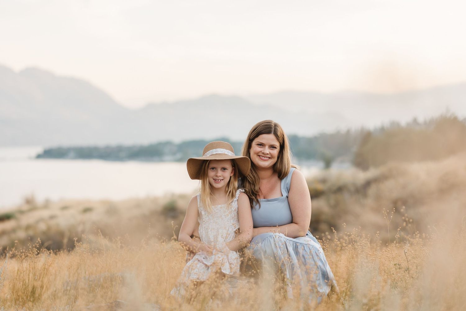A couple sits together in a golden field during sunset as warm light illuminates the mountainous landscape.
