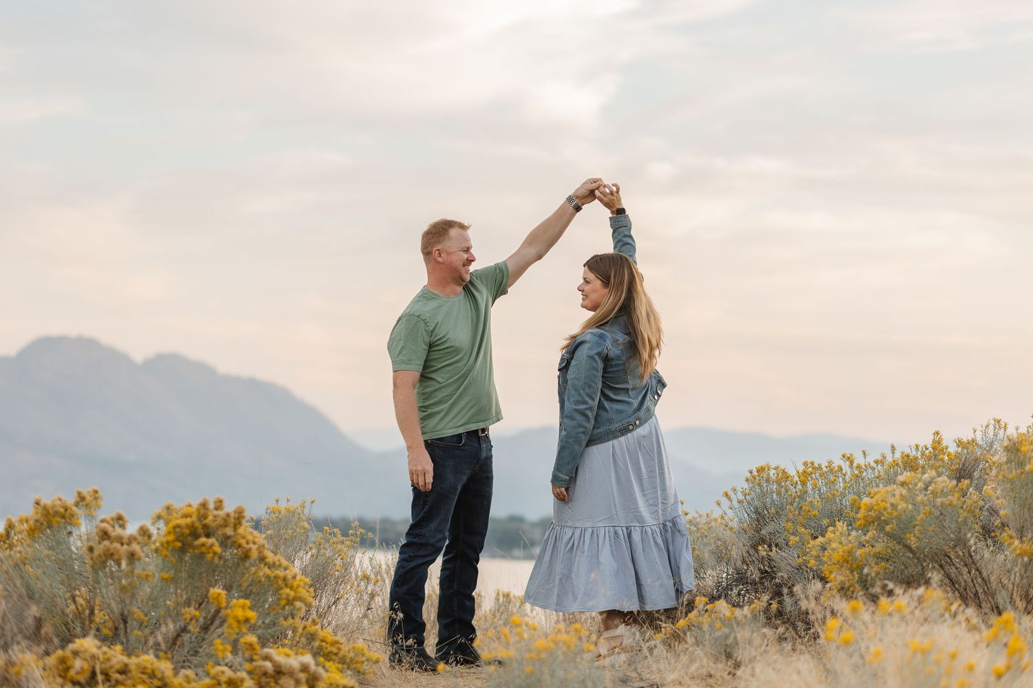 A couple dances together in a field of yellow wildflowers with mountains visible in the background at sunset.