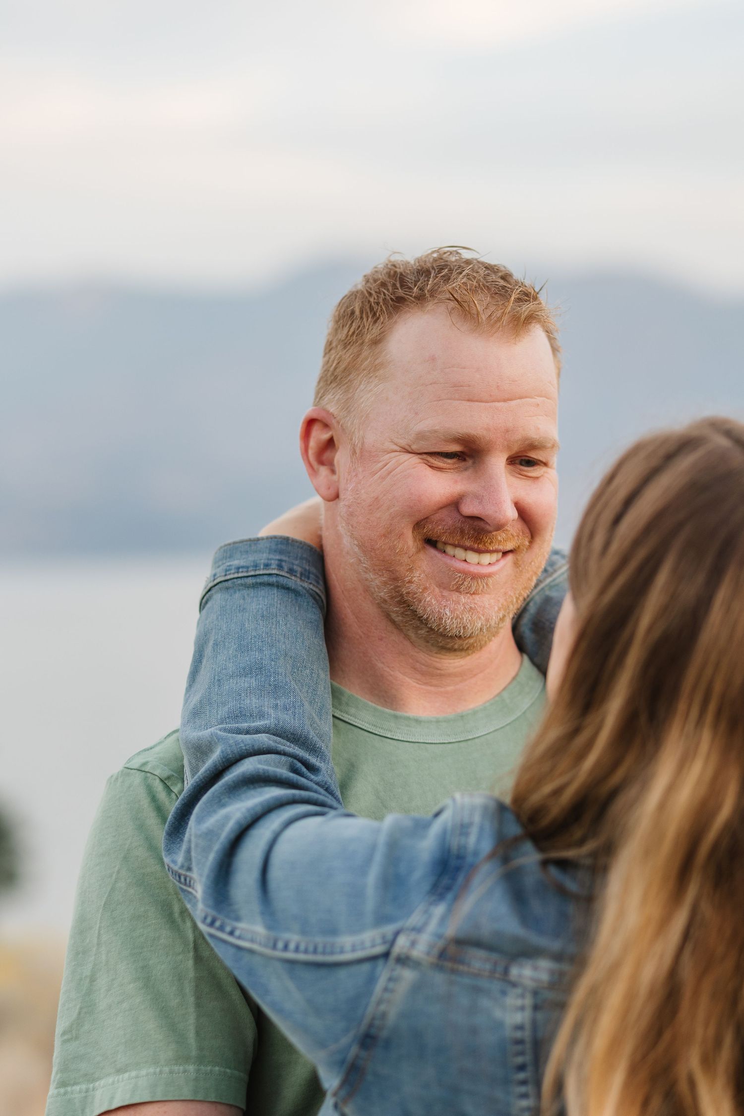 A couple shares a tender moment together outdoors with mountains and water in the background.