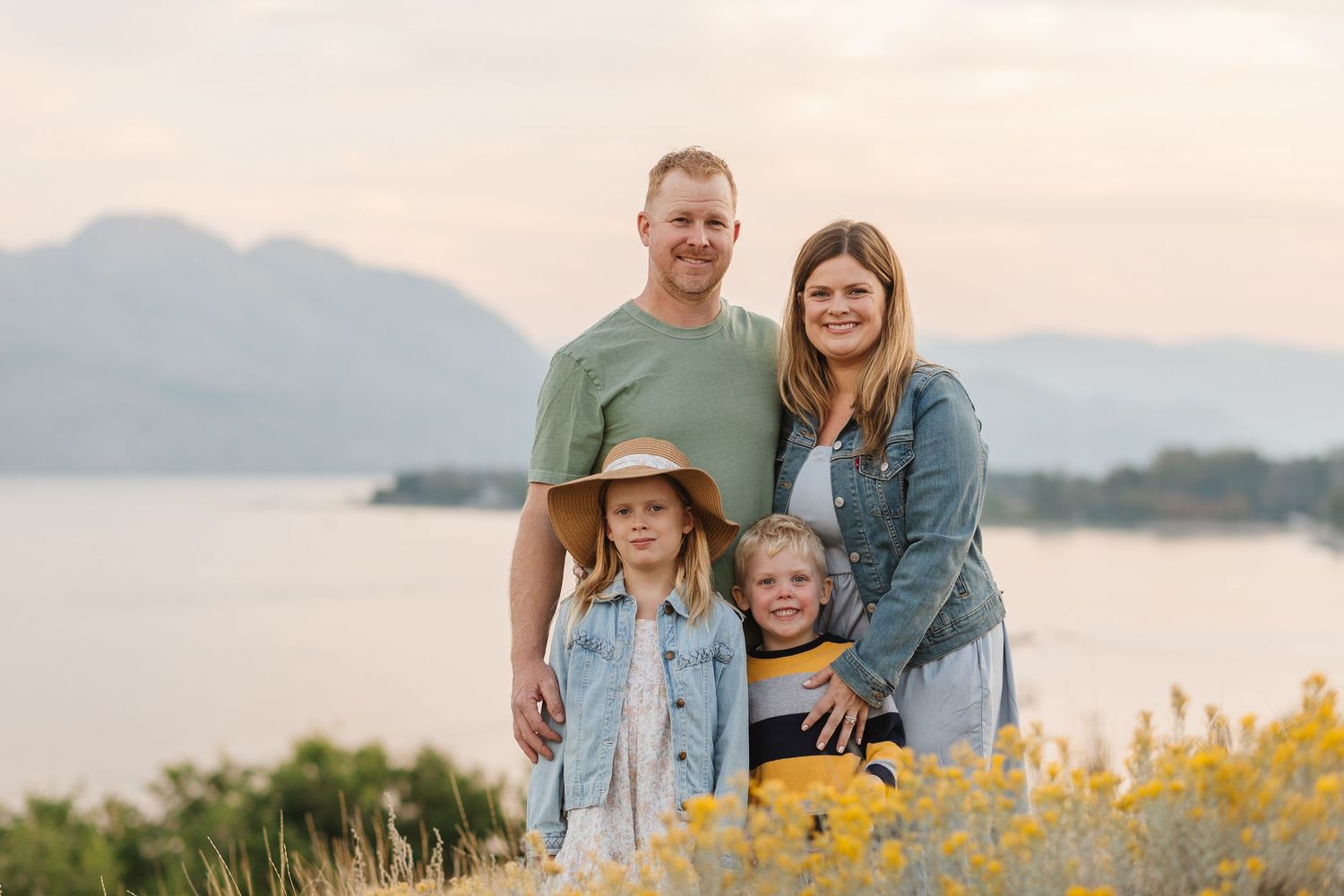 A family stands together on a hilltop overlooking a lake at sunset with yellow wildflowers in the foreground.