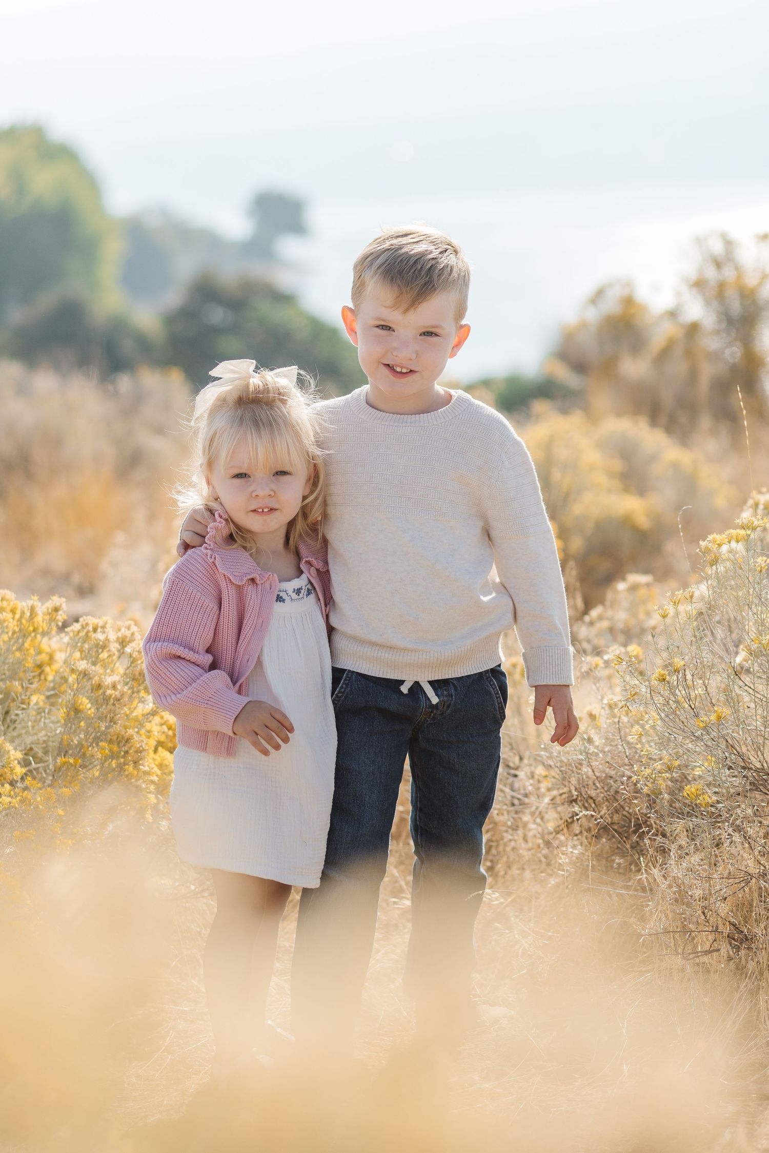 Two young children embrace while standing together in a sunlit field of golden dried grass on a warm autumn day.