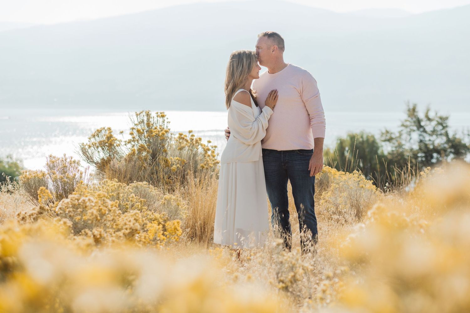 A couple shares an intimate moment on a hilltop overlooking the ocean, surrounded by golden wildflowers at sunset.