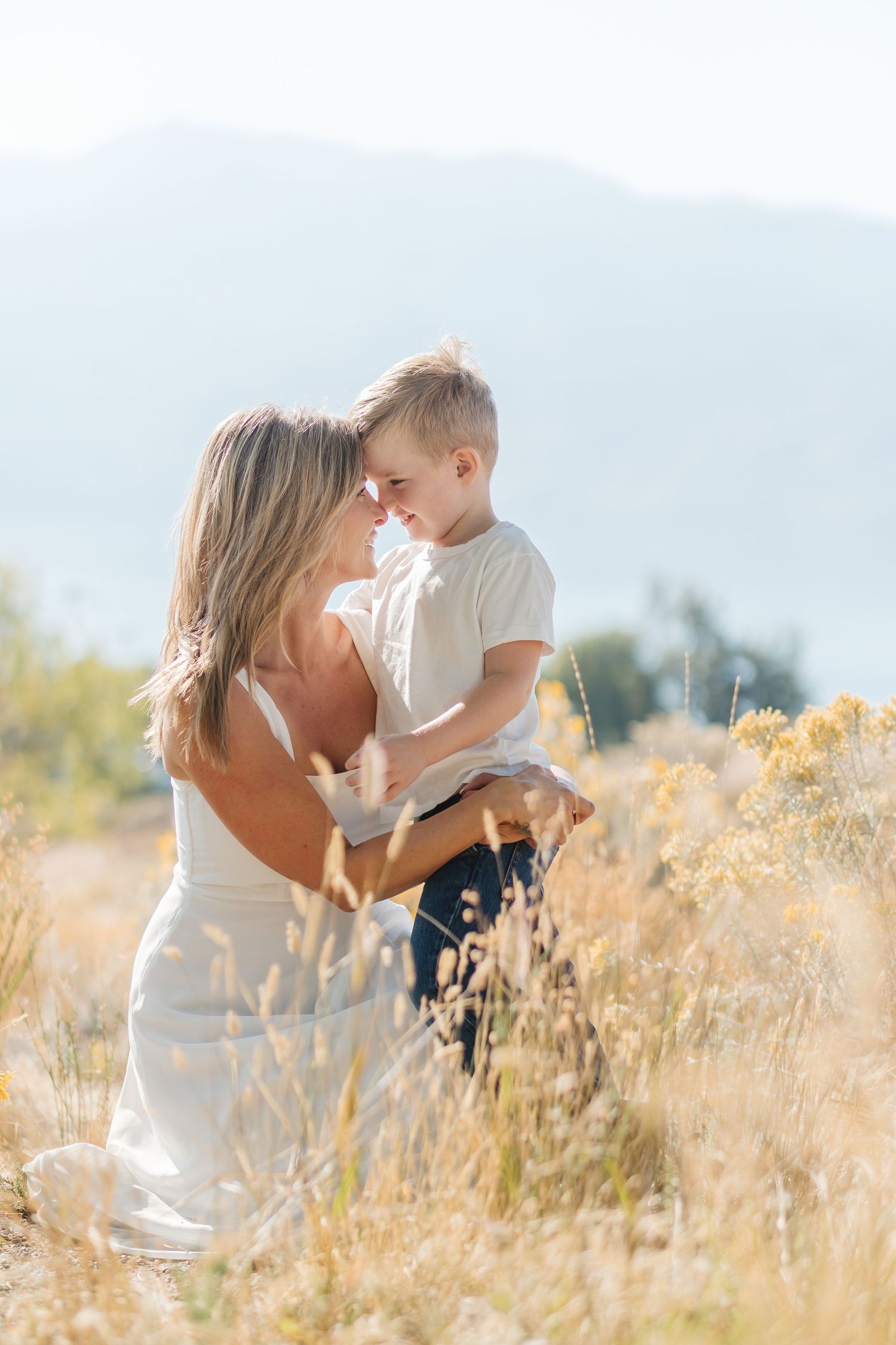 A tender moment shared between parent and child in a sunlit field of golden grass with mountains in the background.