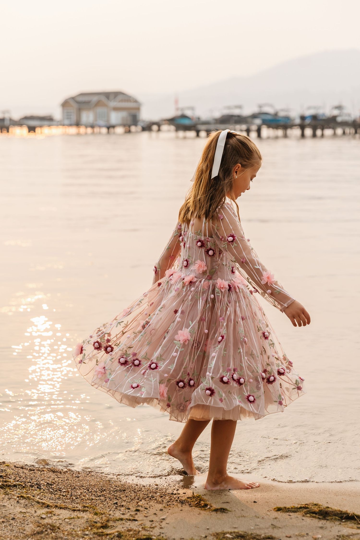 A young child in a pink floral dress twirls and plays along the beach shoreline at sunset.
