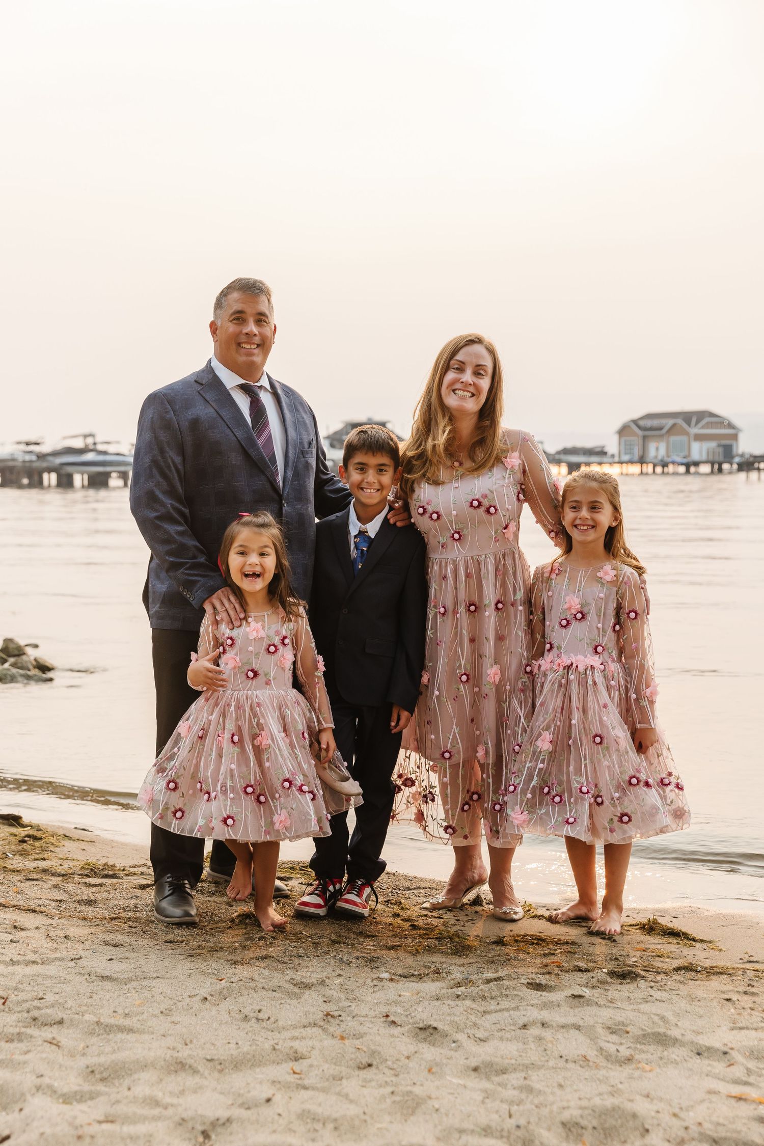 A family in coordinated outfits poses together on a sandy beach with coastal homes visible in the background.