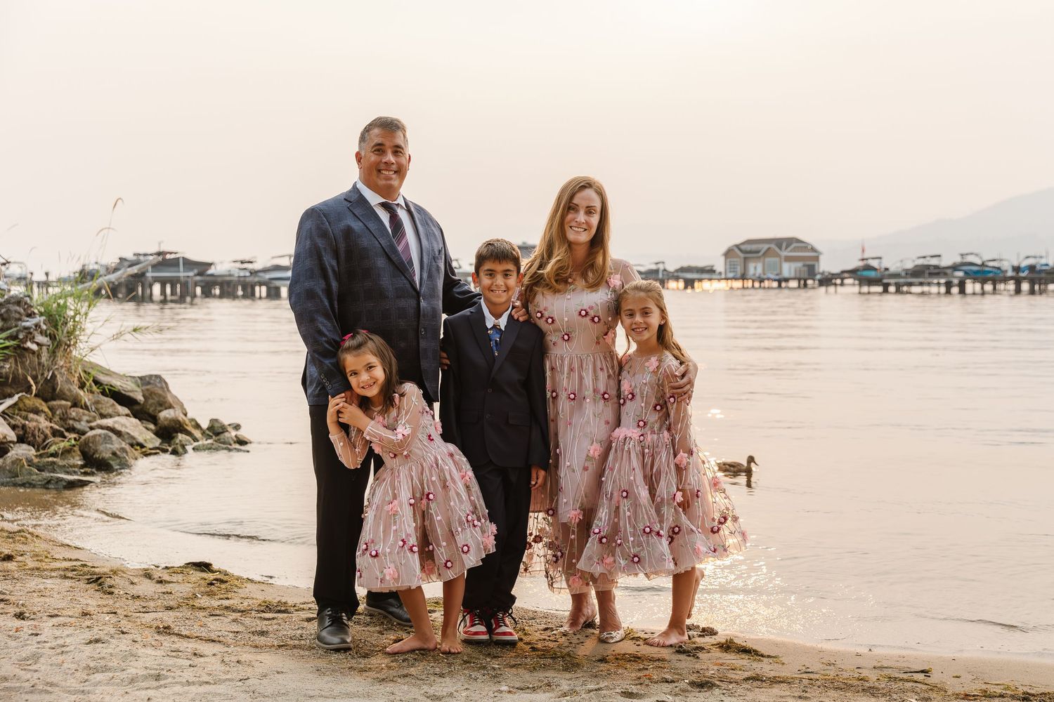 A family in formal attire poses together on a beach at sunset with the ocean in the background.