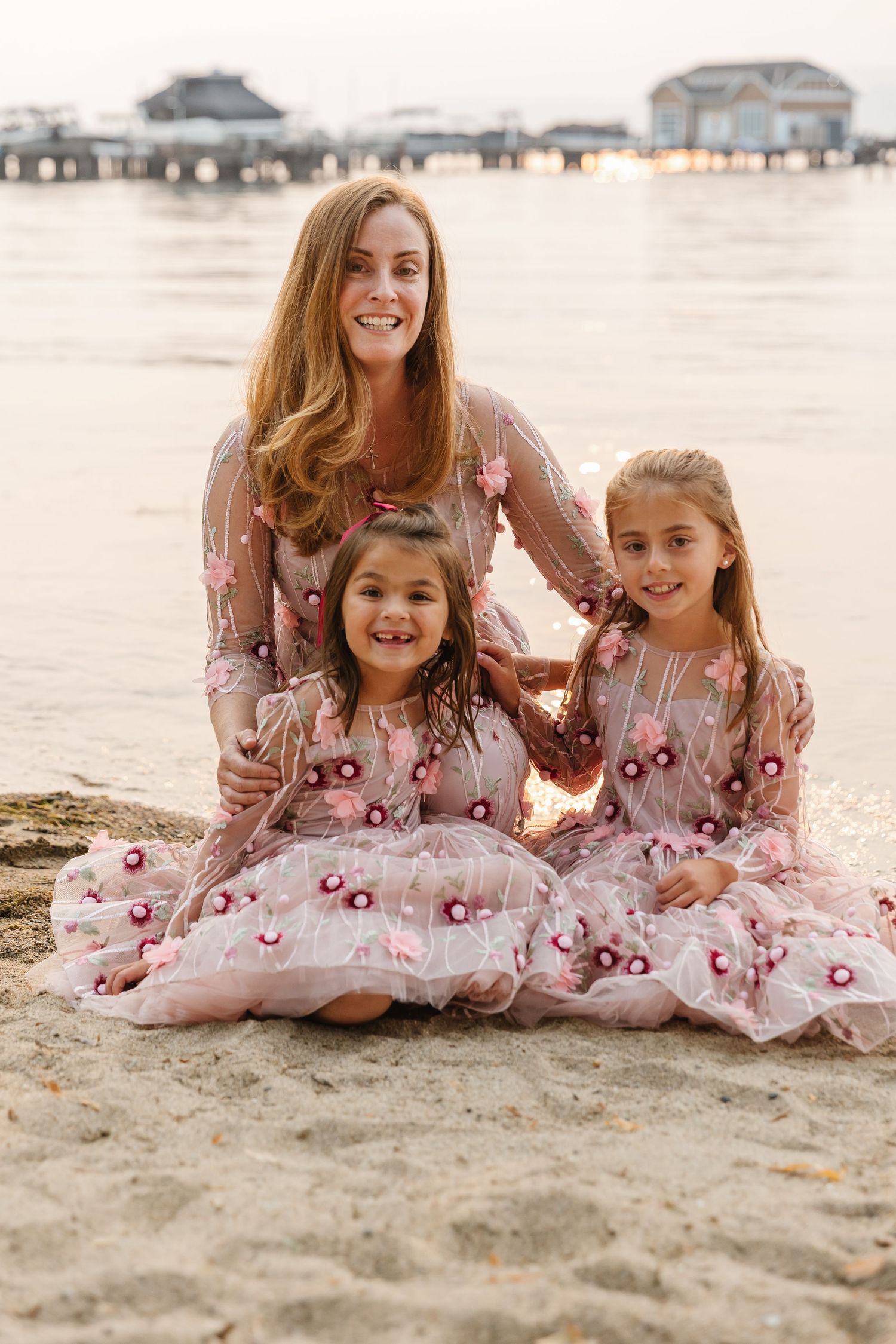 A family wearing matching pink floral dresses poses together on a sandy beach with a pier in the background.