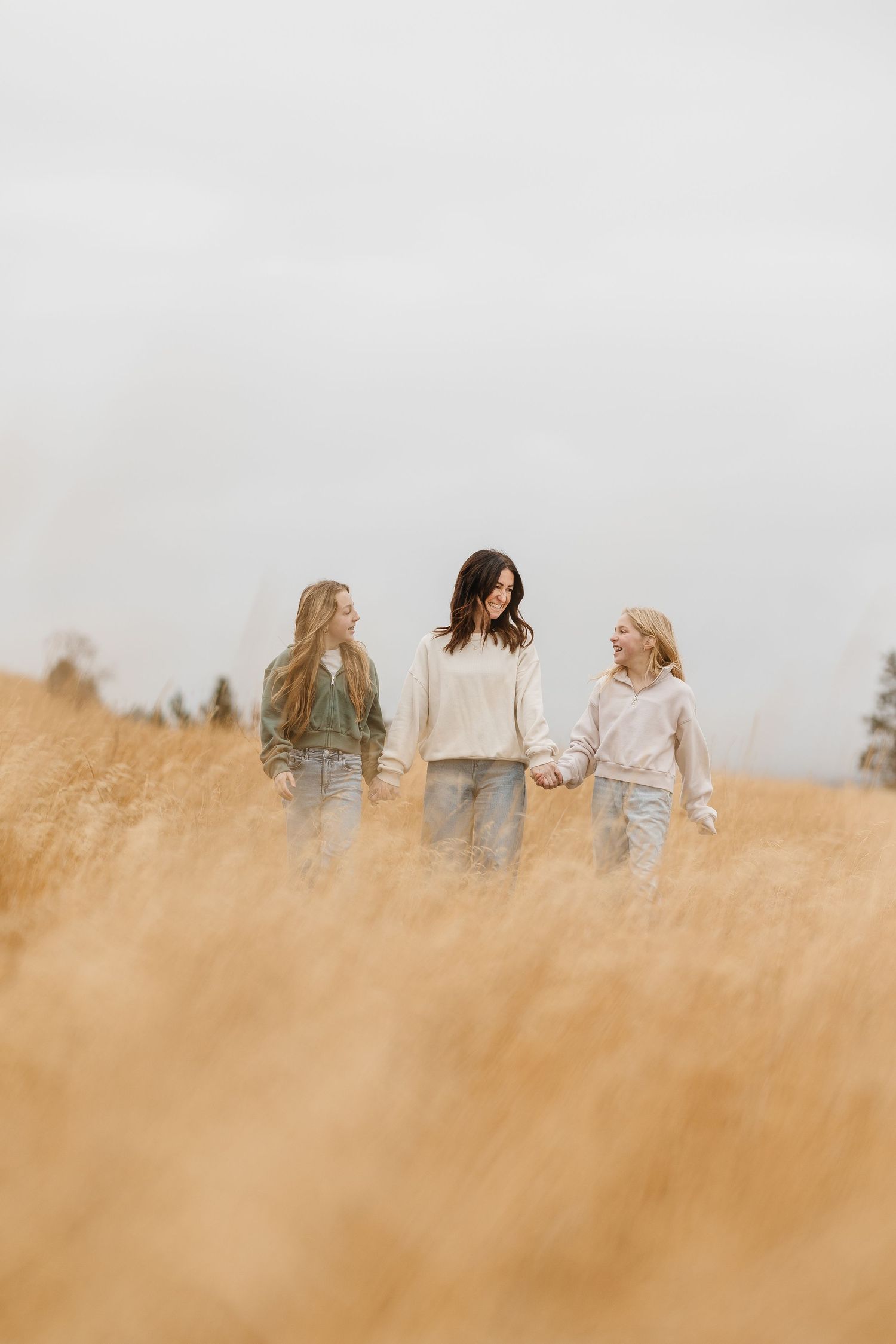 Three people walk hand in hand through tall golden wheat against an overcast sky.