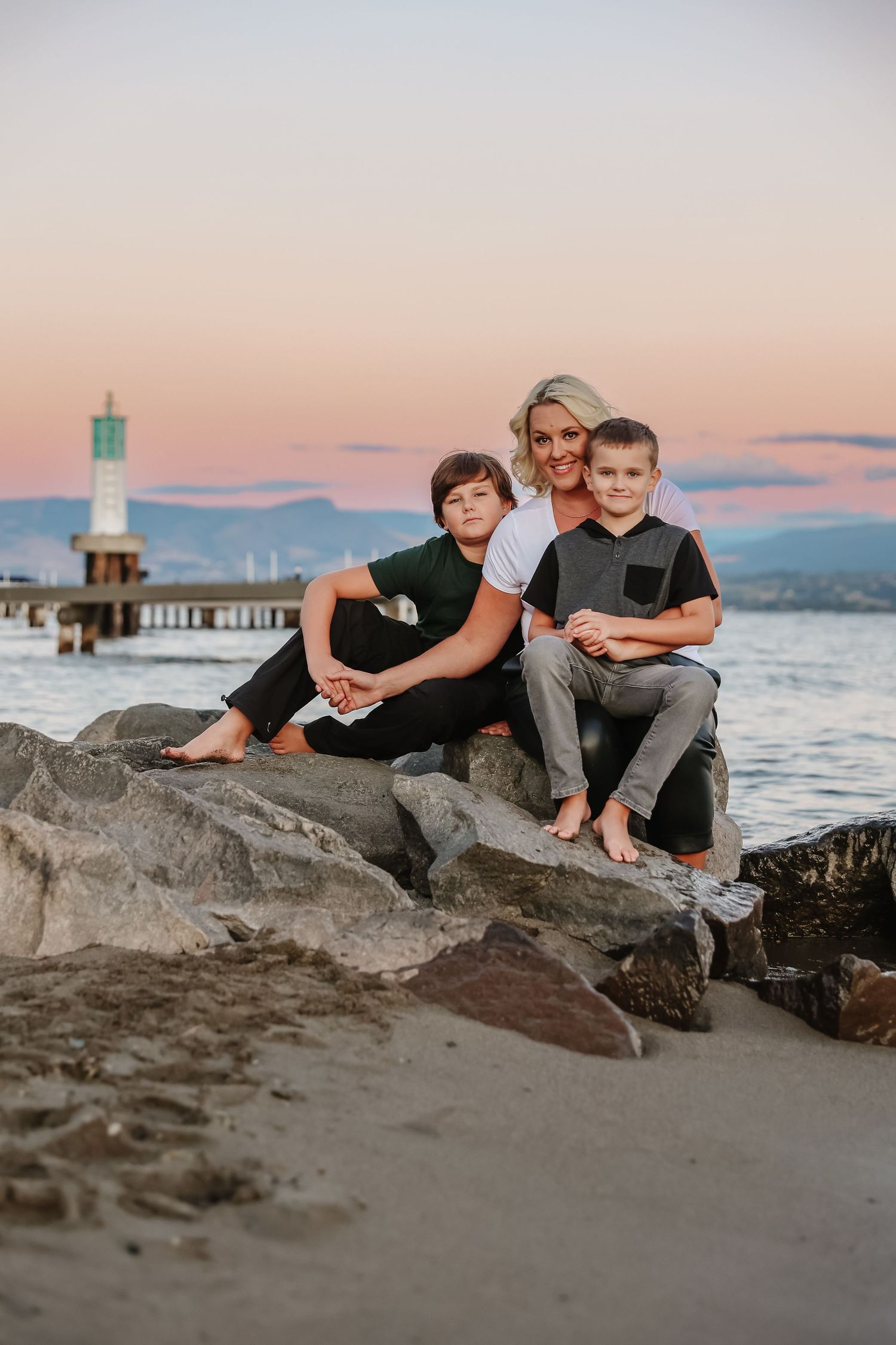 A family portrait on rocky beach with lighthouse and sunset sky in background in west Kelowna in the summer