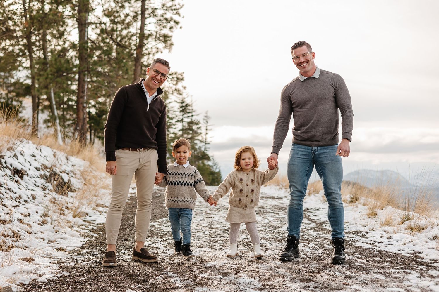 Family of four poses together on snowy mountain trail with evergreen trees in background in Kelowna