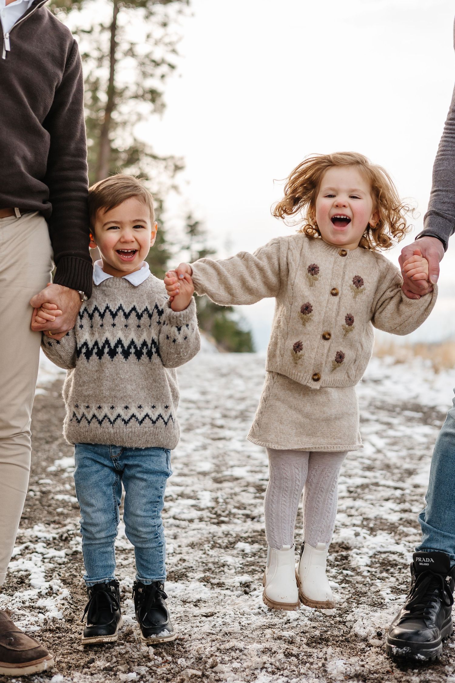 Children wearing matching sweaters hold hands while playing in the snow with mountain views in the background for a fun family photoshoot