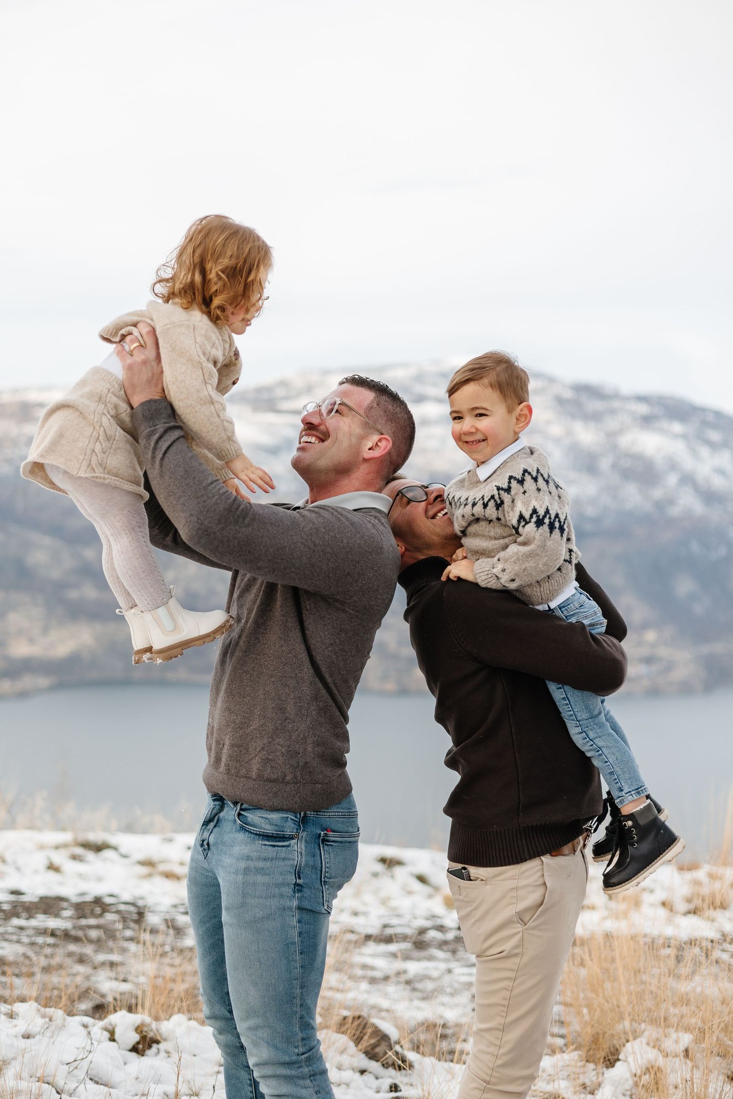Family enjoying a fun moment together on a snowy hillside overlooking a mountain lake for a family photoshoot in kelowna