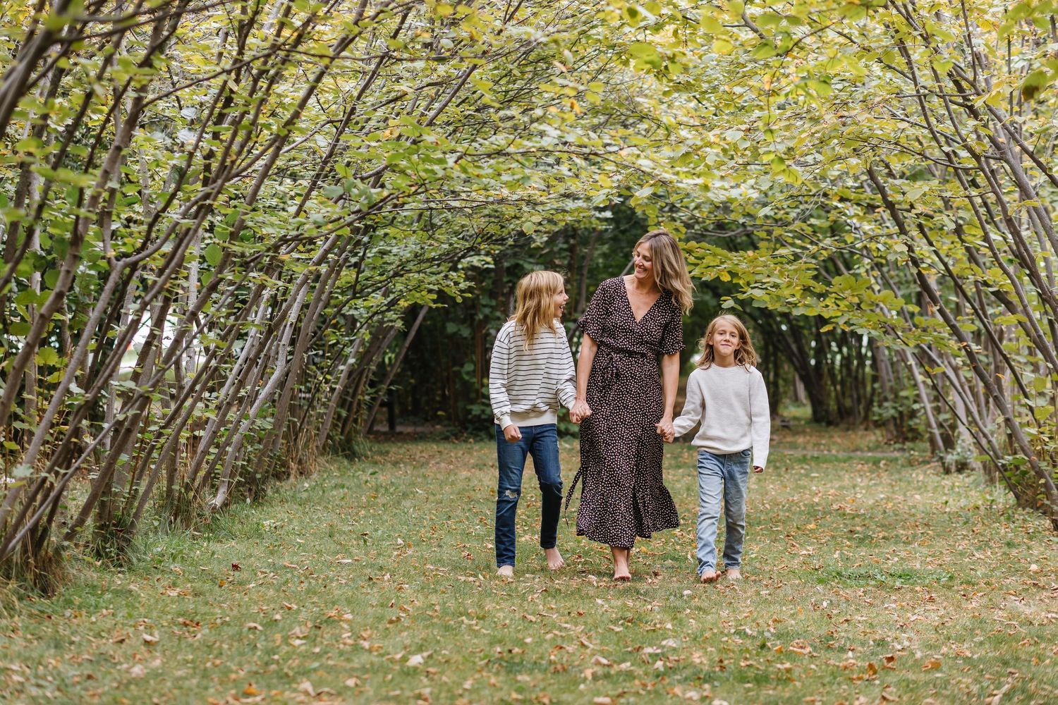 Three people walk together on a grass path beneath an arching canopy of autumn trees.