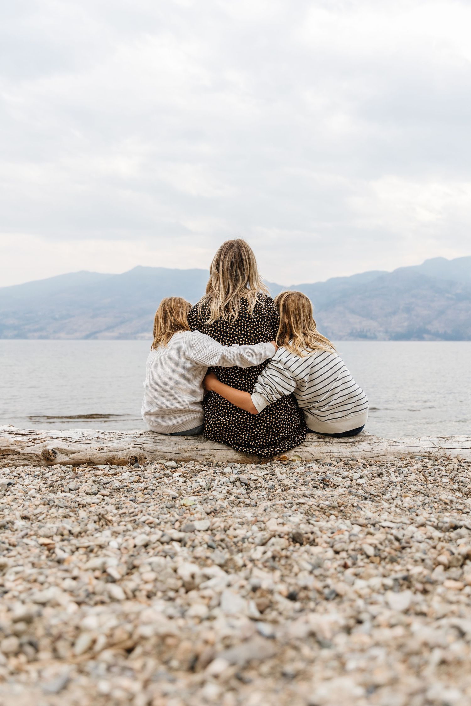 Three friends sit close together on a rocky beach overlooking a serene mountain lake on a cloudy day.
