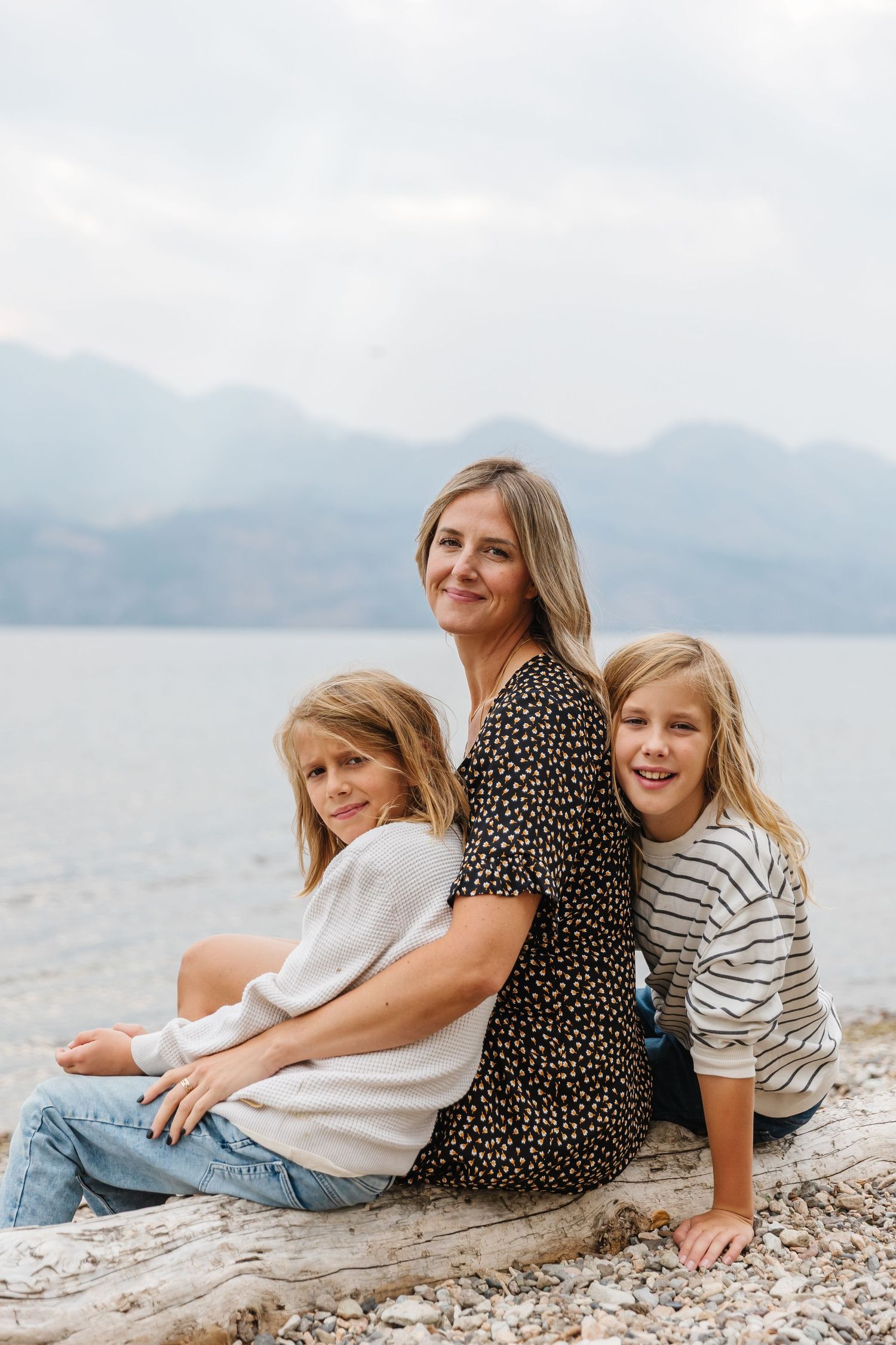 Three people laying on a rocky beach near a lake with mountains in the background on a cloudy day.