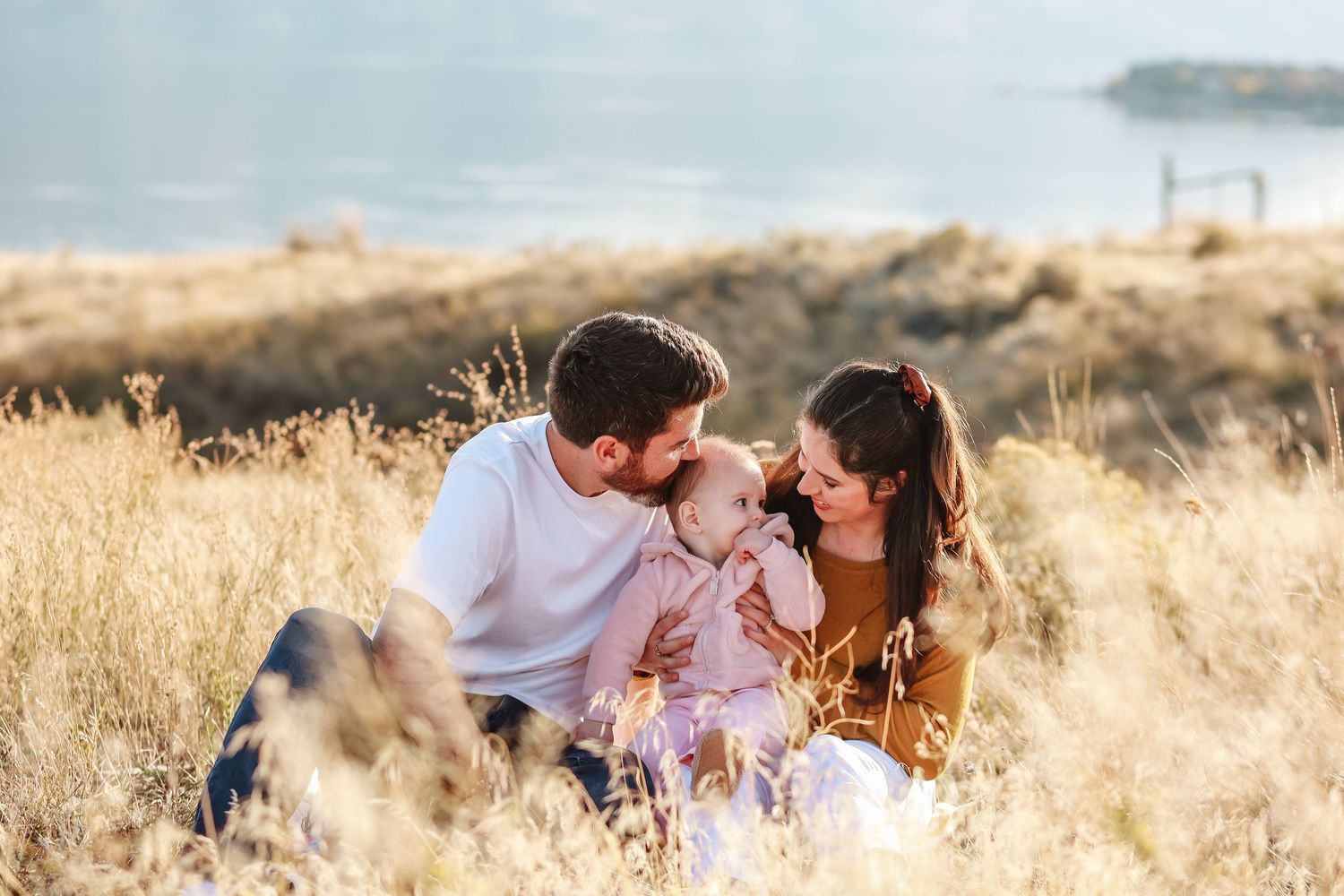 A sweet family moment in a golden field with mountains and water in the distance in West Kelowna at sanctuary gardens