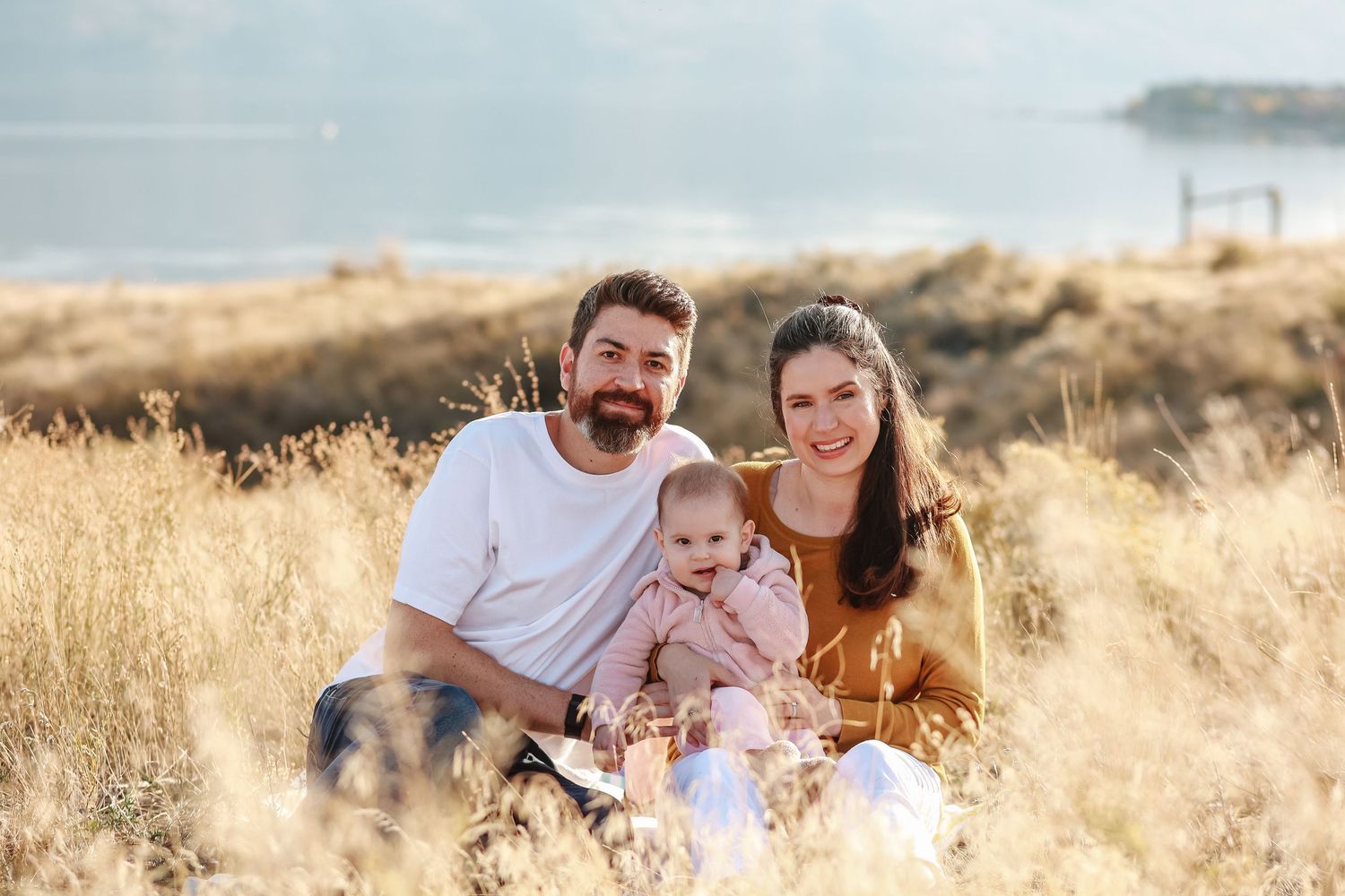 A family portrait in tall grass with water and mountains visible in the background of okanagan lake in West Kelowna at sanctuary gardens