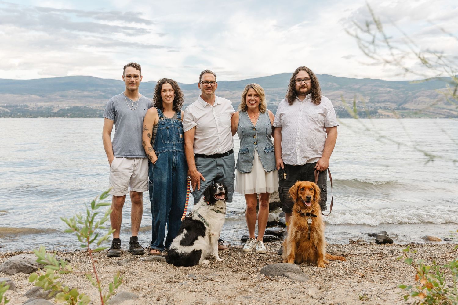 A group sits with their dogs on a rocky beach overlooking a scenic mountain lake on a cloudy day.