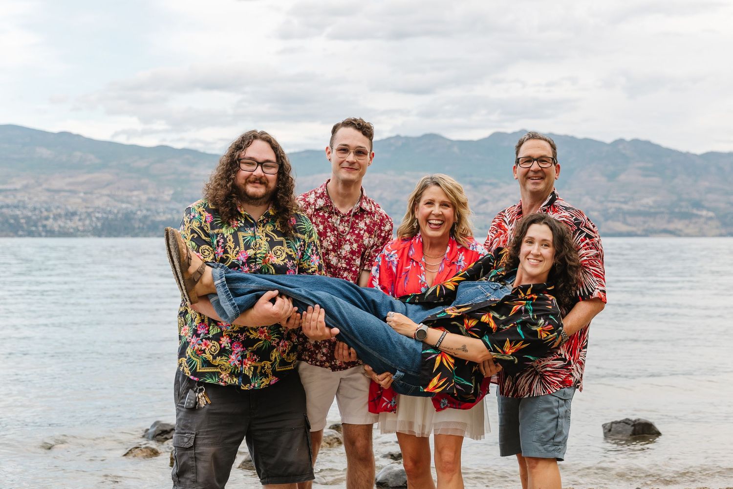 A group of friends lift and carry someone playfully at a scenic lakeside location with mountains in the background.