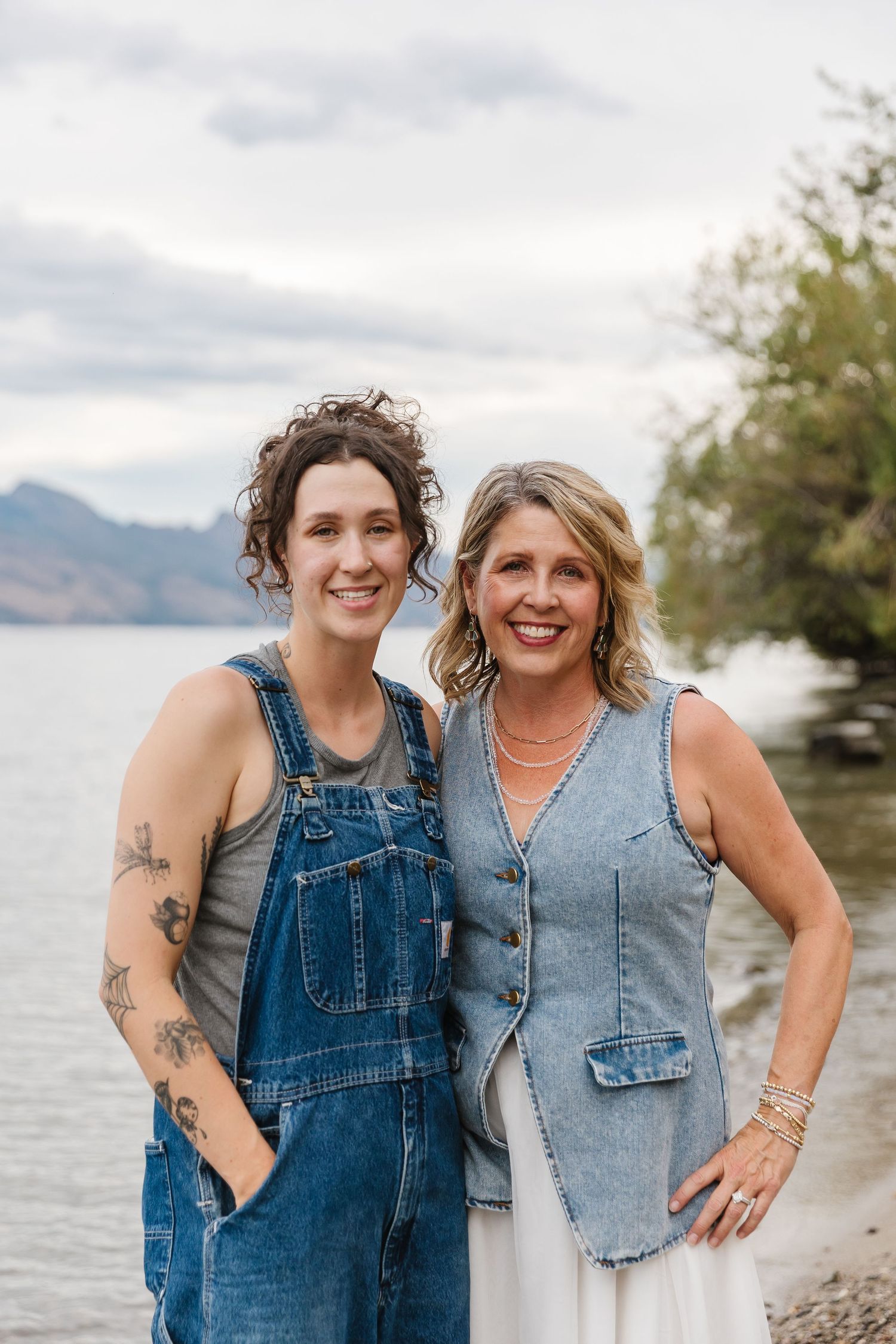 Two people wearing denim outfits smile and embrace while posing together on a scenic beach with mountains in background.