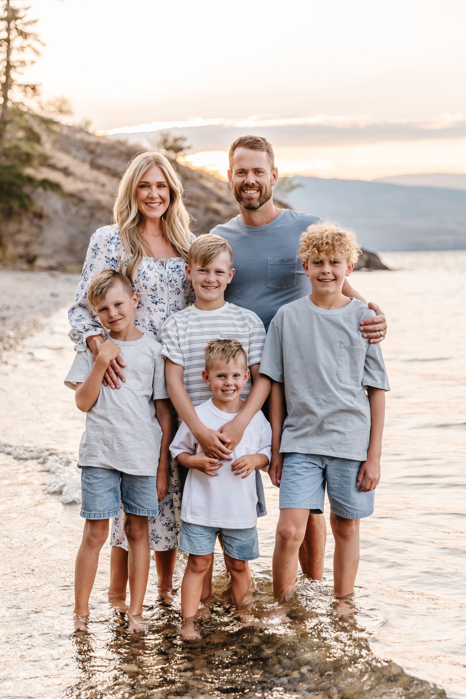 Family wearing coordinated blue and white outfits standing in shallow water at sunset during a photoshoot in kelowna at Bertram creek park