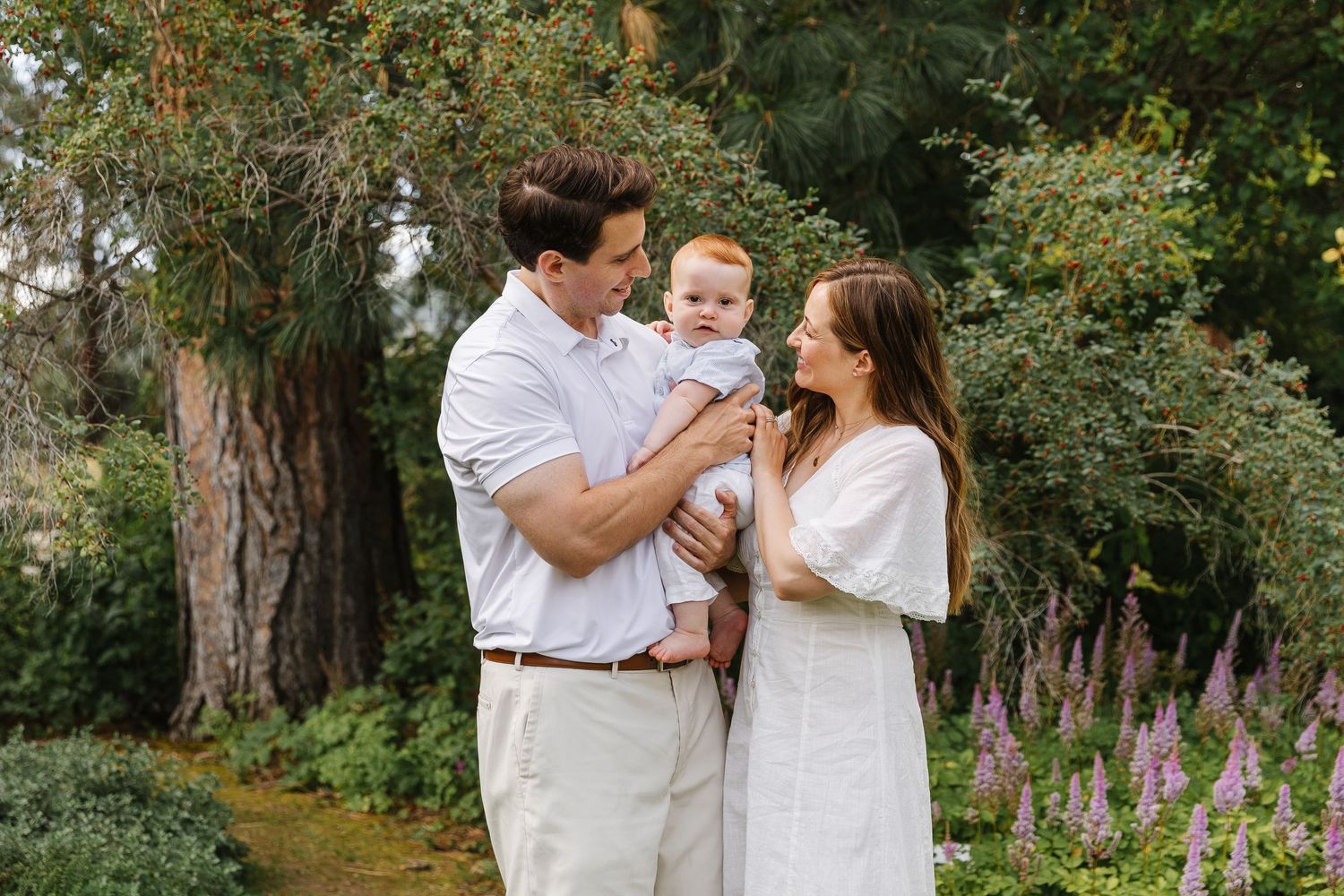 A family wearing white clothing poses together in a garden setting with purple wildflowers blooming in the background.