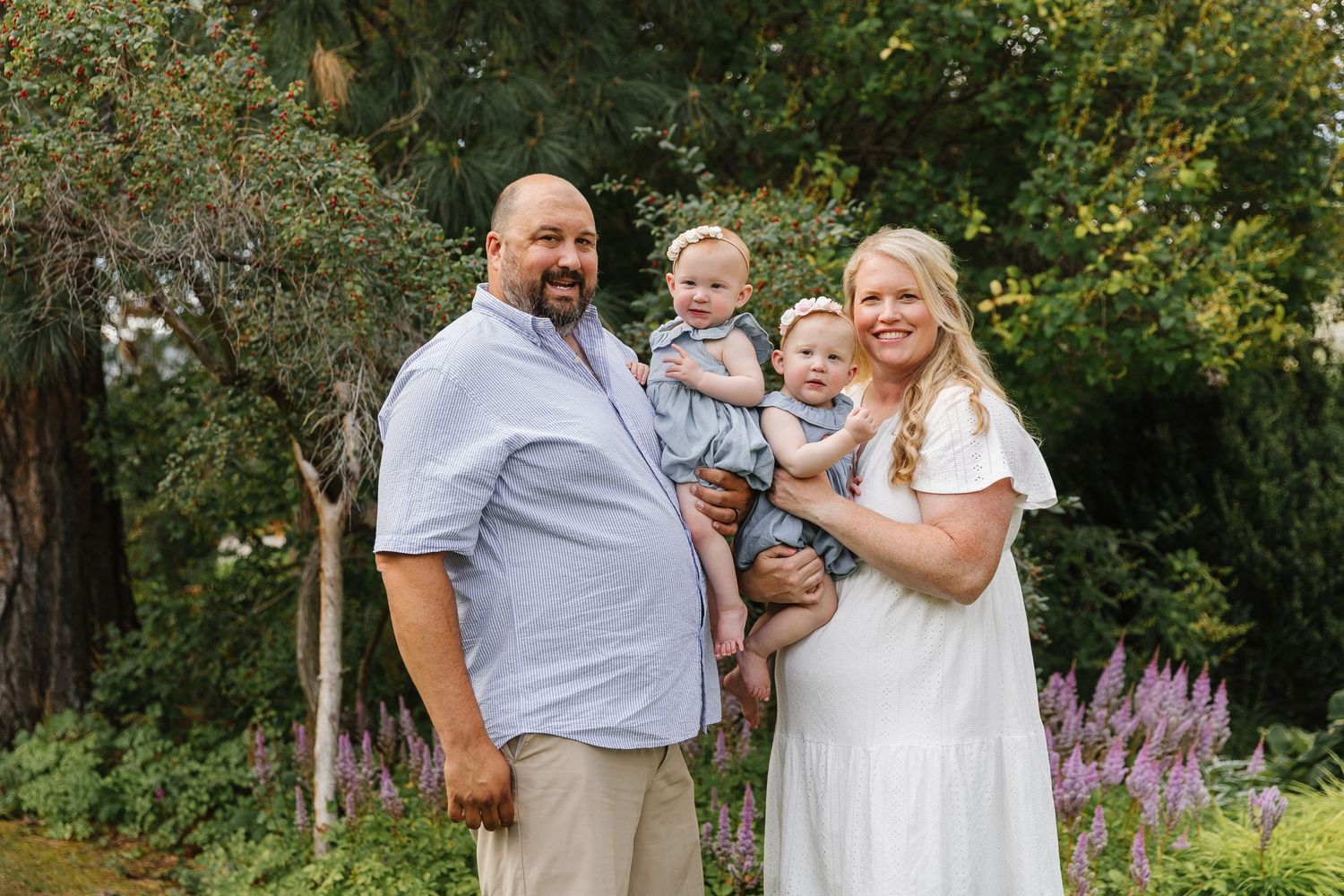 A family wearing coordinated white and gray outfits poses together in a lush garden setting during a photo session.