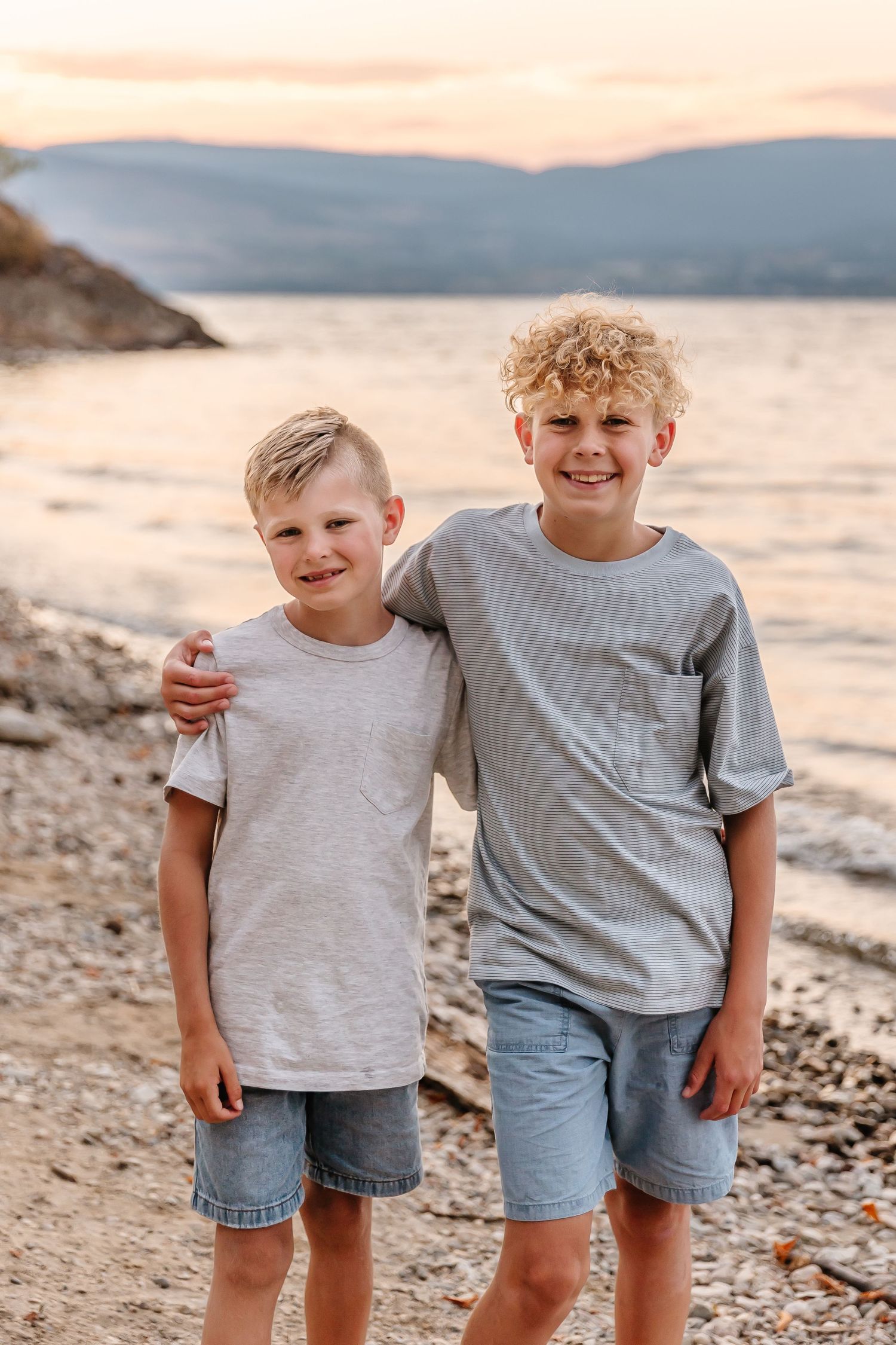 Two children in casual summer clothing standing together on a rocky beach at sunset during a family photoshoot in kelowna at Bertram park