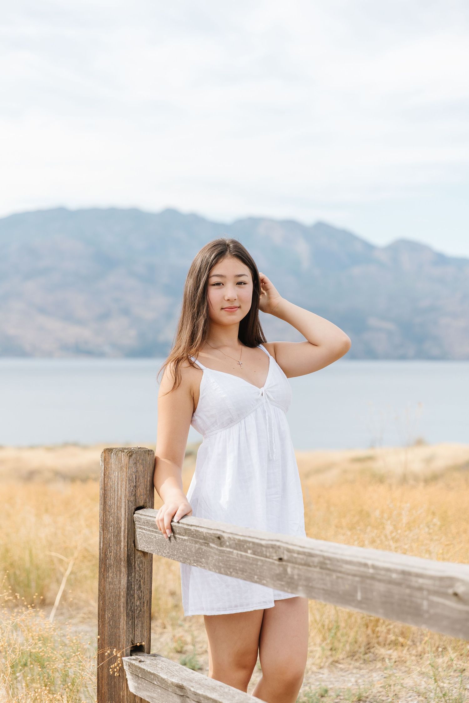 A person in a white dress poses by a wooden fence with mountains and water in the background.