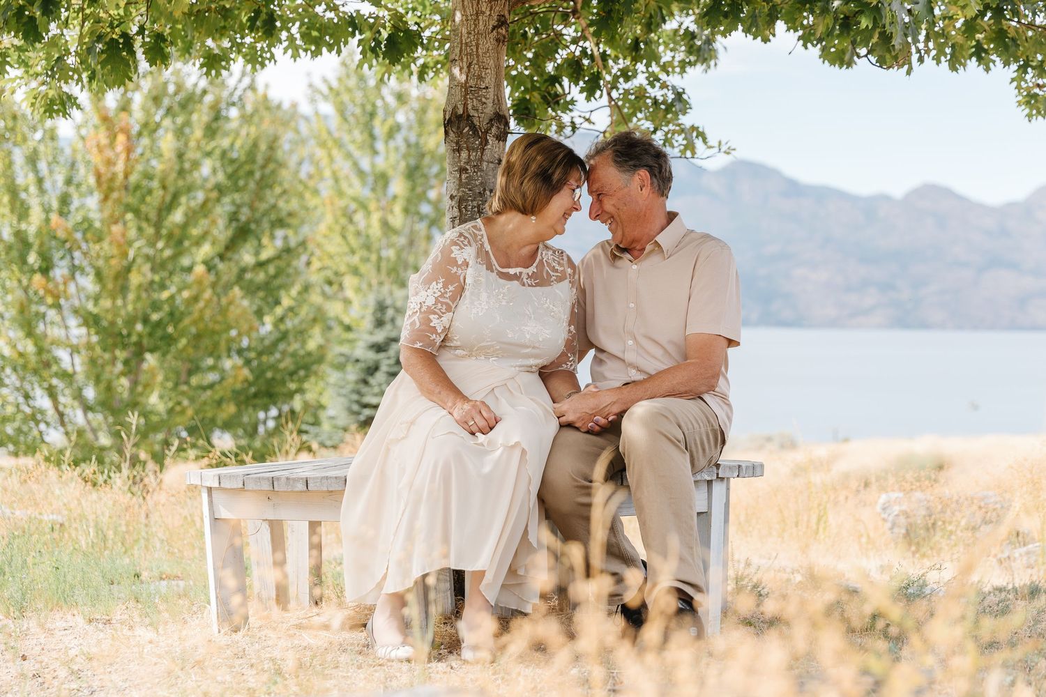 A couple sits together on a wooden bench overlooking a serene lake and mountain landscape on a sunny day.