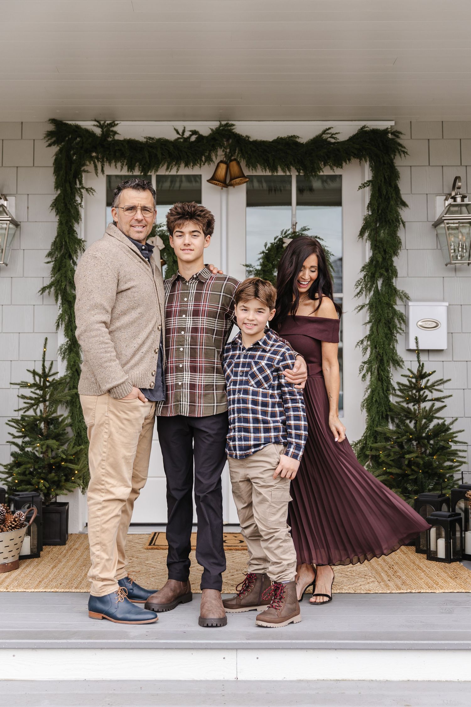 Family wearing coordinated plaid and burgundy outfits pose on a front porch decorated with holiday greenery.