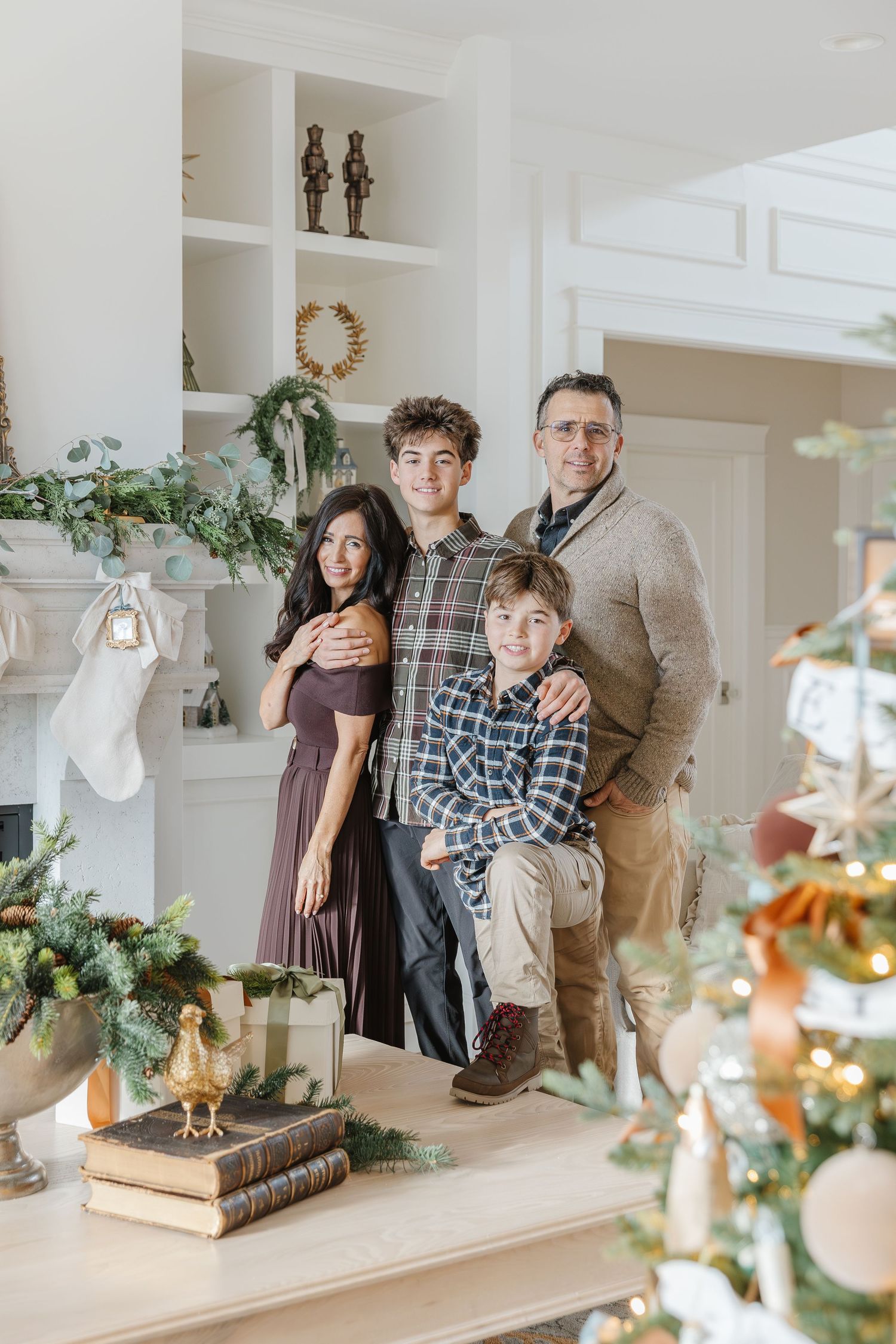 A family stands together near white built-in shelves decorated with holiday greenery and decor in a bright interior space.
