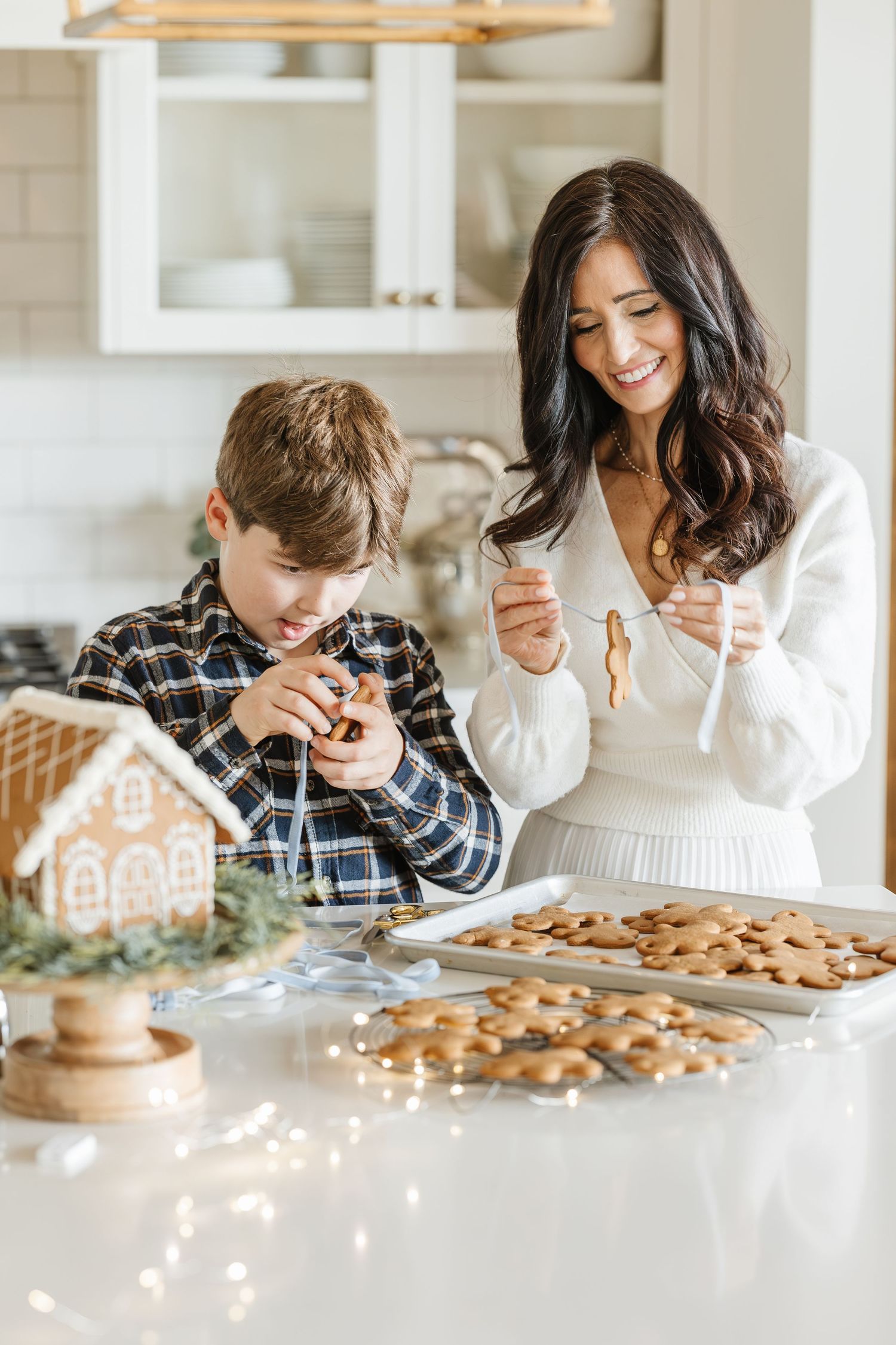 Mother and son decorating gingerbread cookies together in bright white kitchen with gingerbread house nearby.
