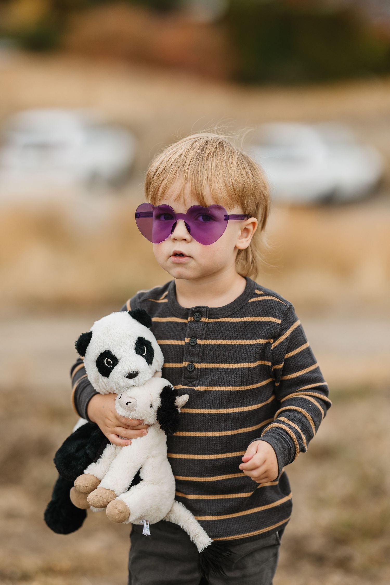 A small child in sunglasses holds a plush panda toy while wearing a striped shirt outdoors.