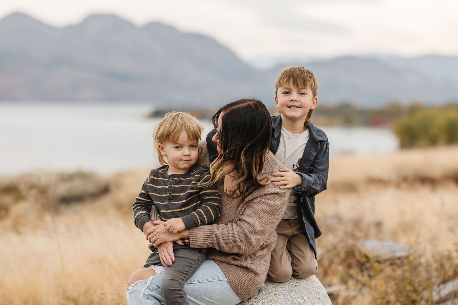 Family shares tender moment together in autumn field with mountains and lake in background.