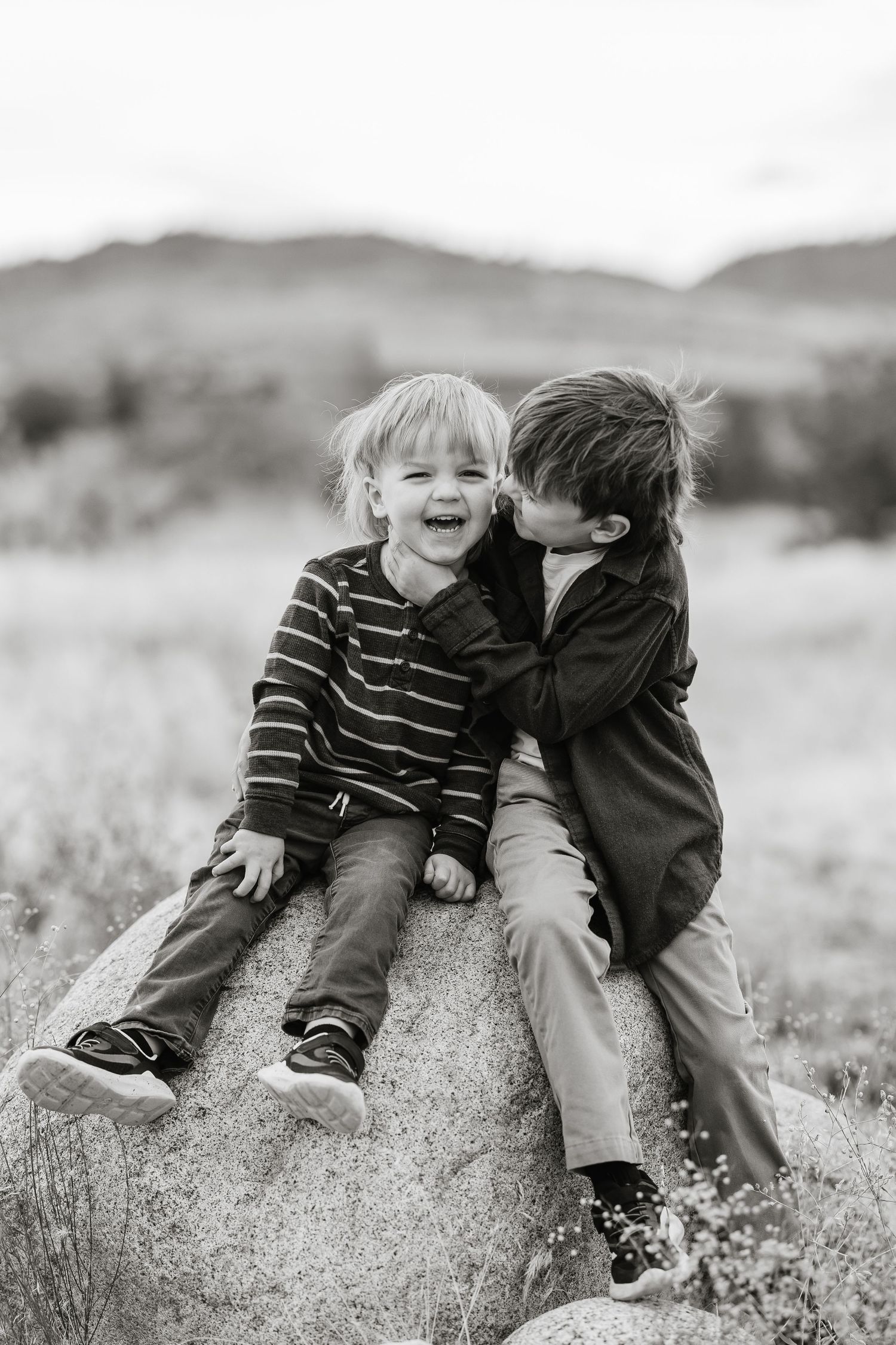 Black and white photo of two children sitting on a rock and sharing a sweet moment together in an outdoor setting.