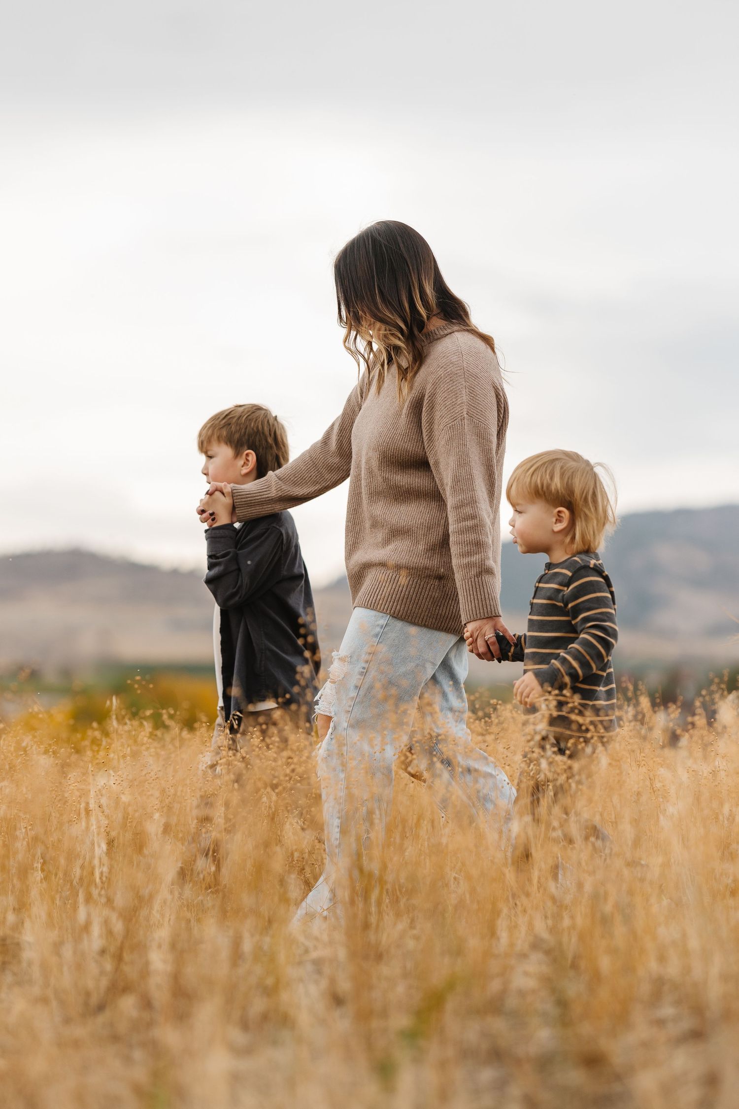 A family walks together through a golden autumn field with mountains in the background on an overcast day.