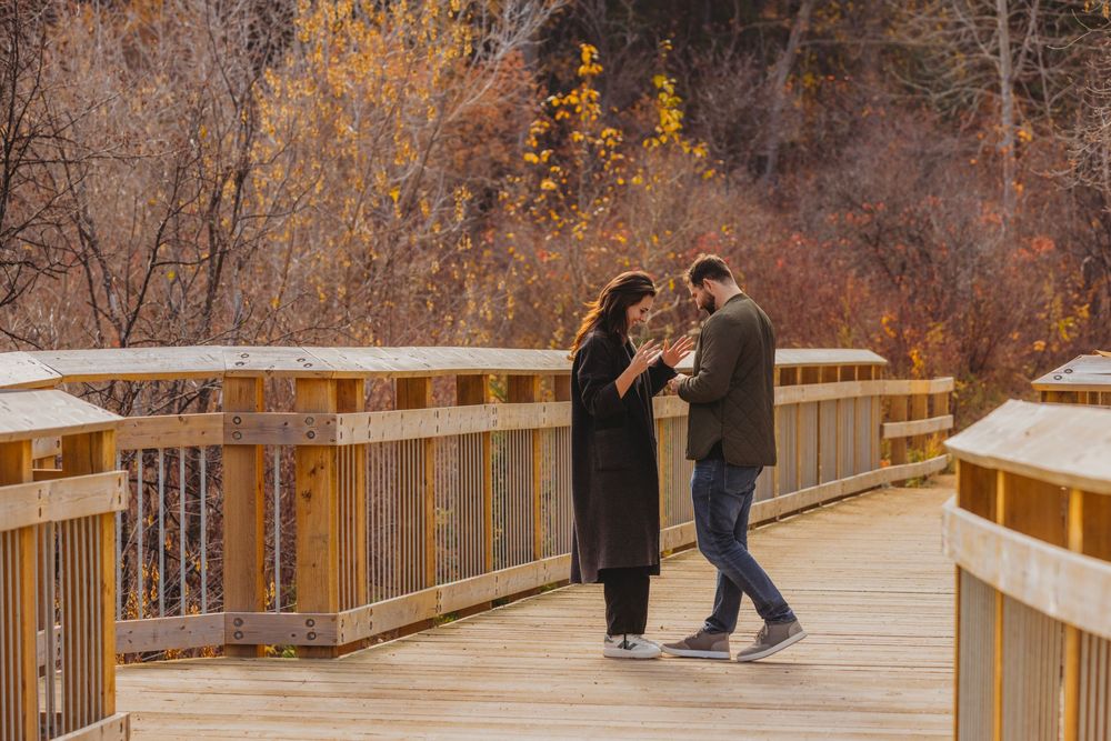 Couple sharing a tender moment on a wooden boardwalk bridge surrounded by autumn foliage in a park.