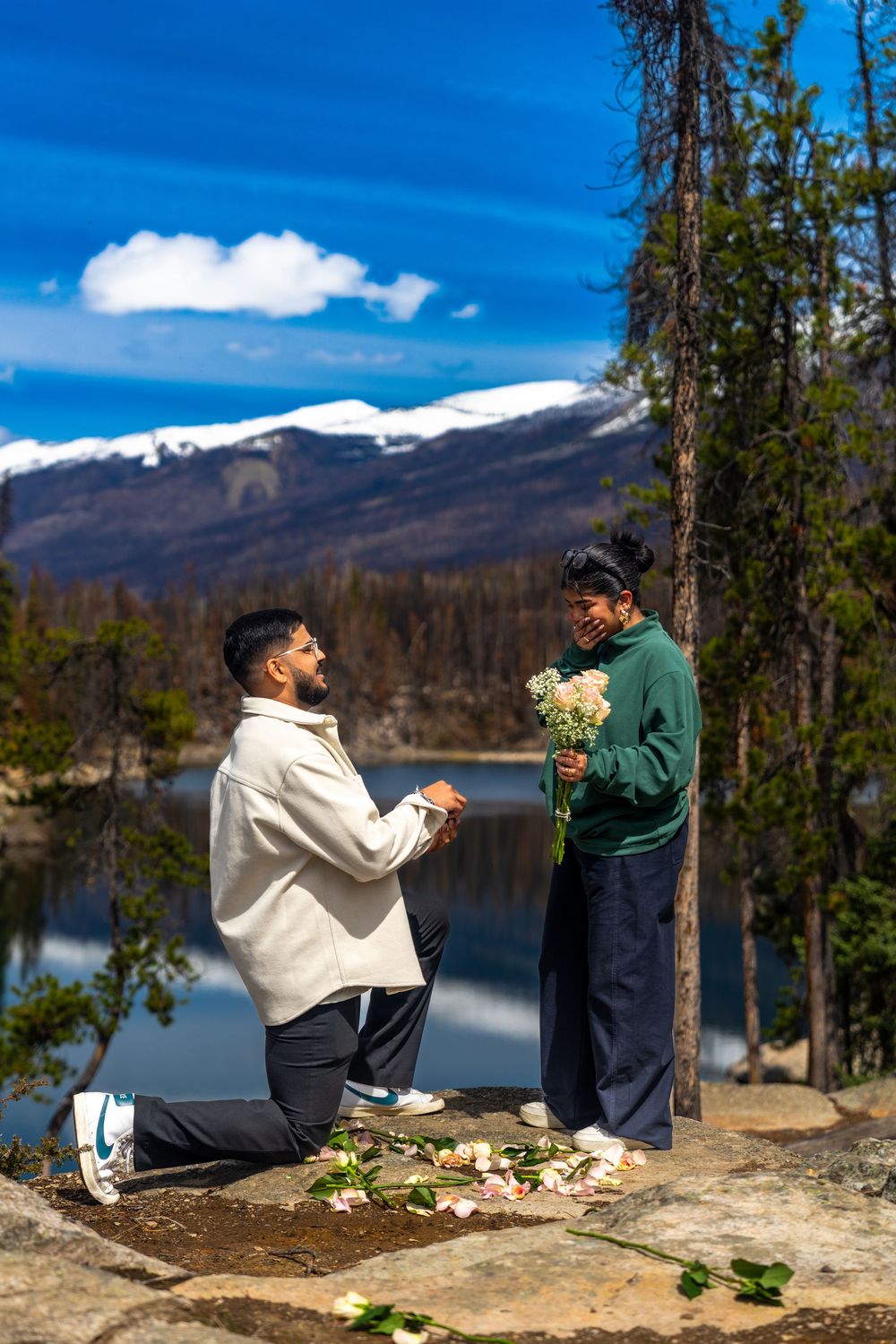 Man proposes to woman on one knee by a mountain lake with snow-capped peaks and blue sky backdrop.