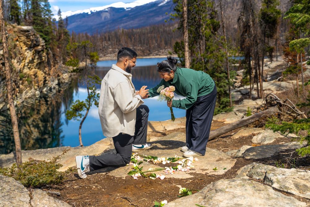 Man proposes to partner on rocky overlook with scenic mountain lake and pine forest backdrop.
