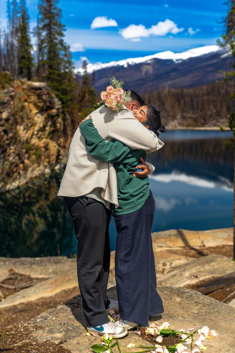 Two people embracing near a mountain lake, one holding a bouquet, with snow-capped peaks and blue sky behind them.
