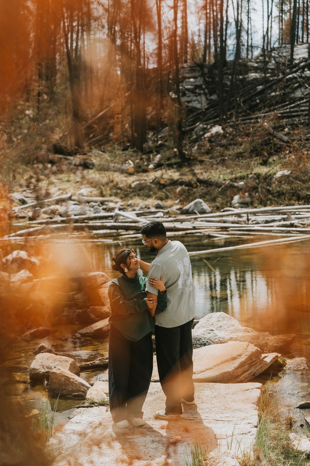 Couple embracing romantically on rocks by a forest stream in warm autumn sunlight.