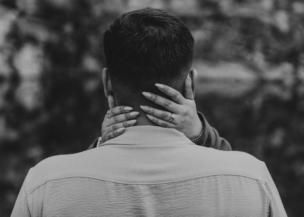 Black and white photo of a man seen from behind, touching the back of his neck with one hand.