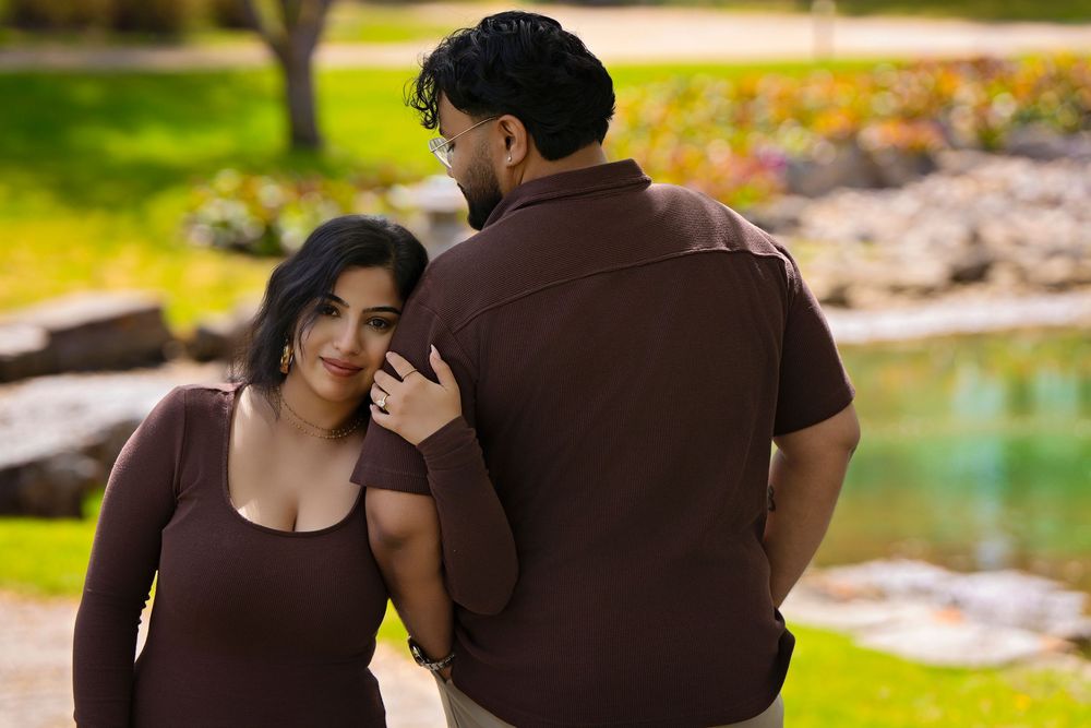 A couple in matching brown outfits poses outdoors near a pond, sharing a tender moment in a lush green park.