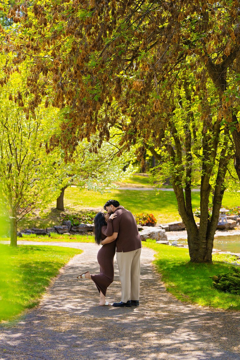 Couple sharing a romantic kiss on a winding garden path surrounded by lush green trees in a scenic park.