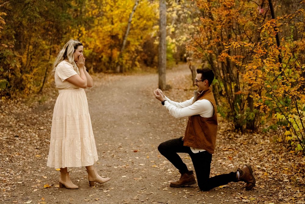 Man proposing on one knee to woman on autumn forest path surrounded by golden fall foliage.