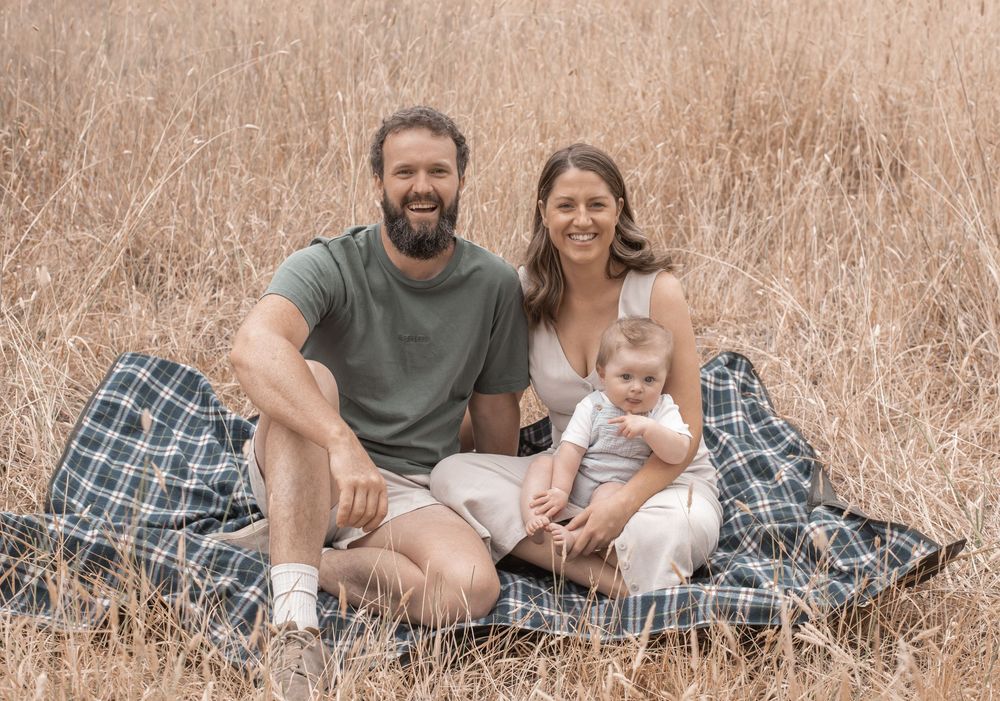 A family sits together on a plaid blanket in a field of tall brown grass on a sunny day.