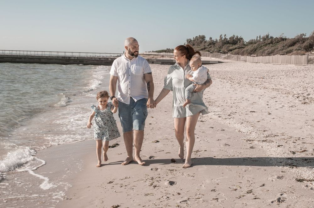 A family walks together along a sunny beach shoreline with waves lapping at their feet.