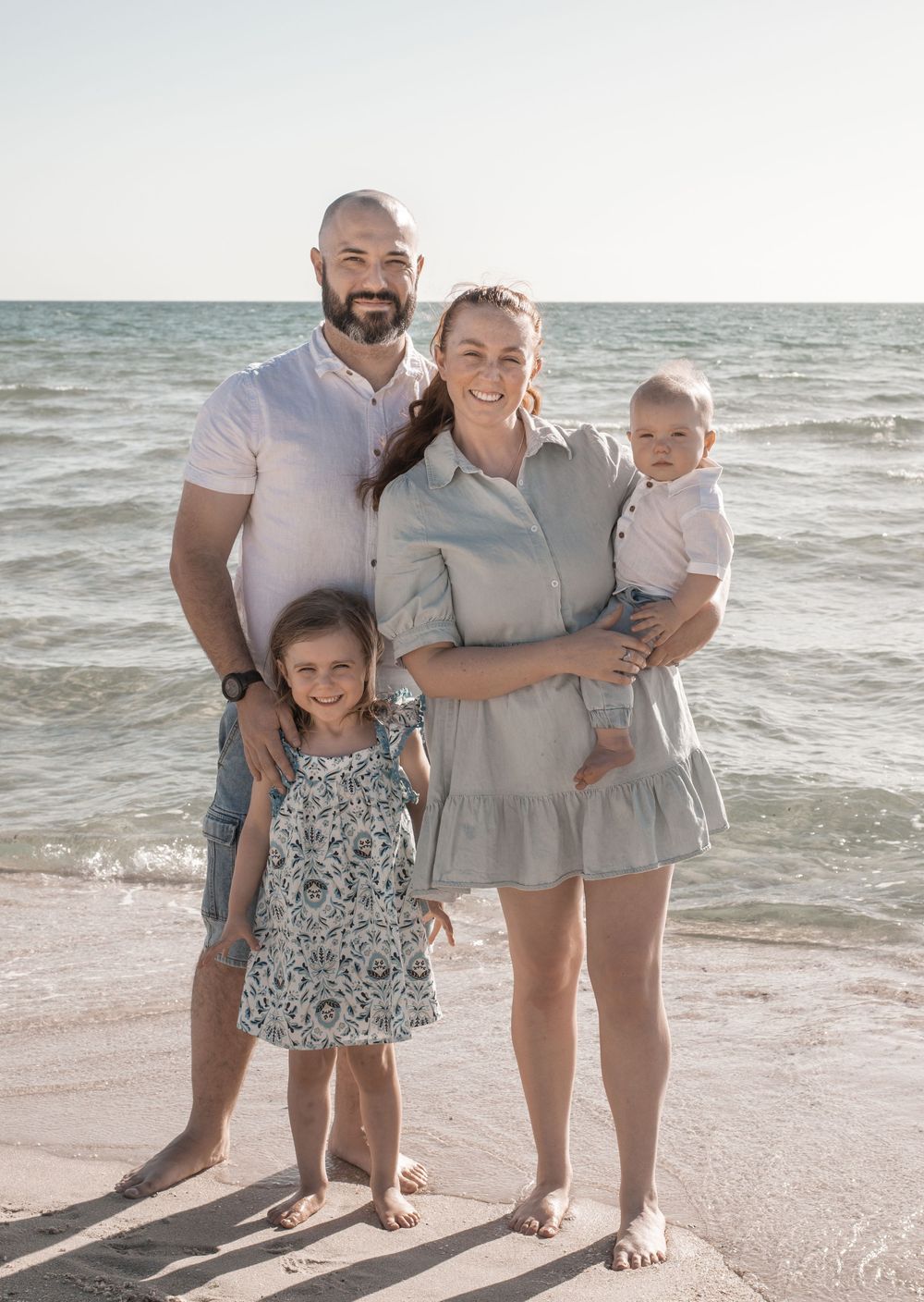 A family of three poses together on a sunny beach with waves crashing in the background.