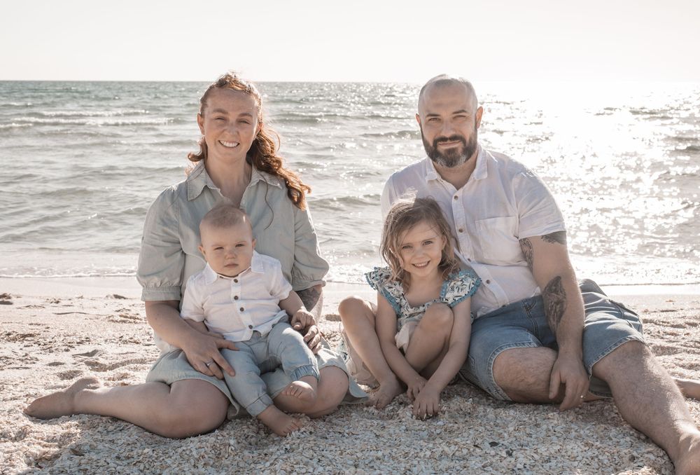 A family wearing coordinated blue and white outfits poses together on a sunny beach at sunset.
